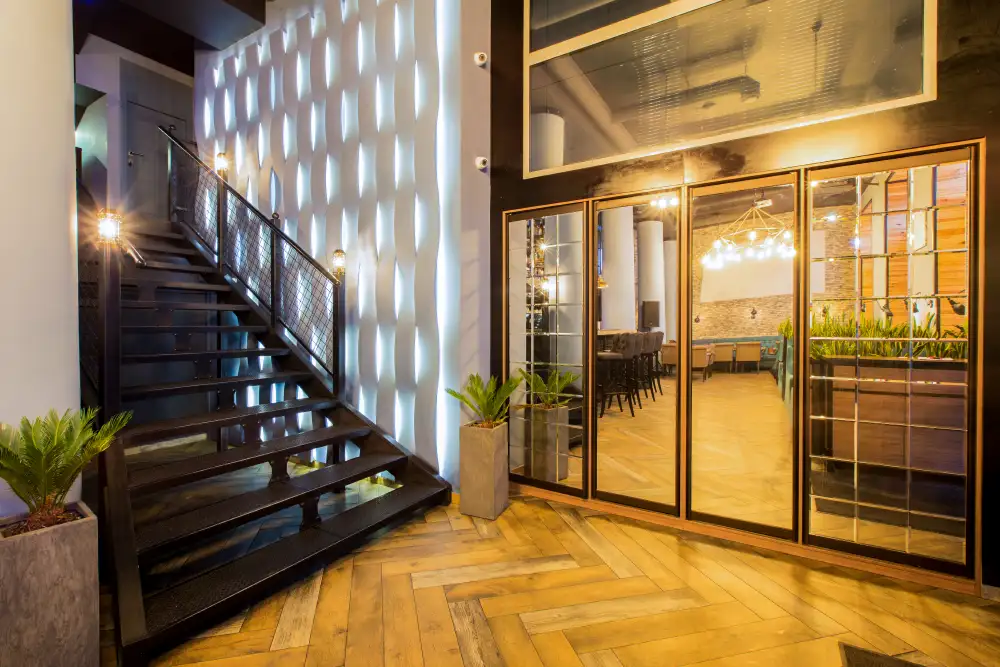 Blick in den hellen, luxuriösen Eingangsbereich eines Restaurants mit einer modernen Holztreppe, einer leuchtenden Wandstruktur und Spiegeln, neutralisiert Gerüche für einen makellosen ersten Eindruck.